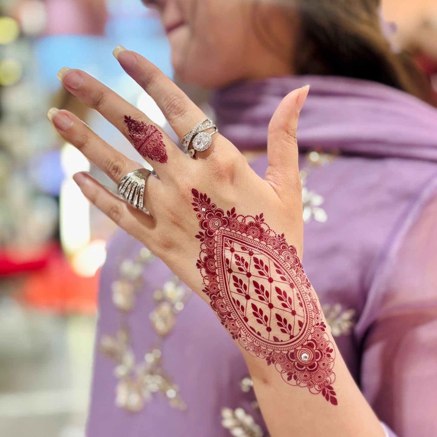 Hand with intricate red henna design, wearing multiple rings, against a blurred background.