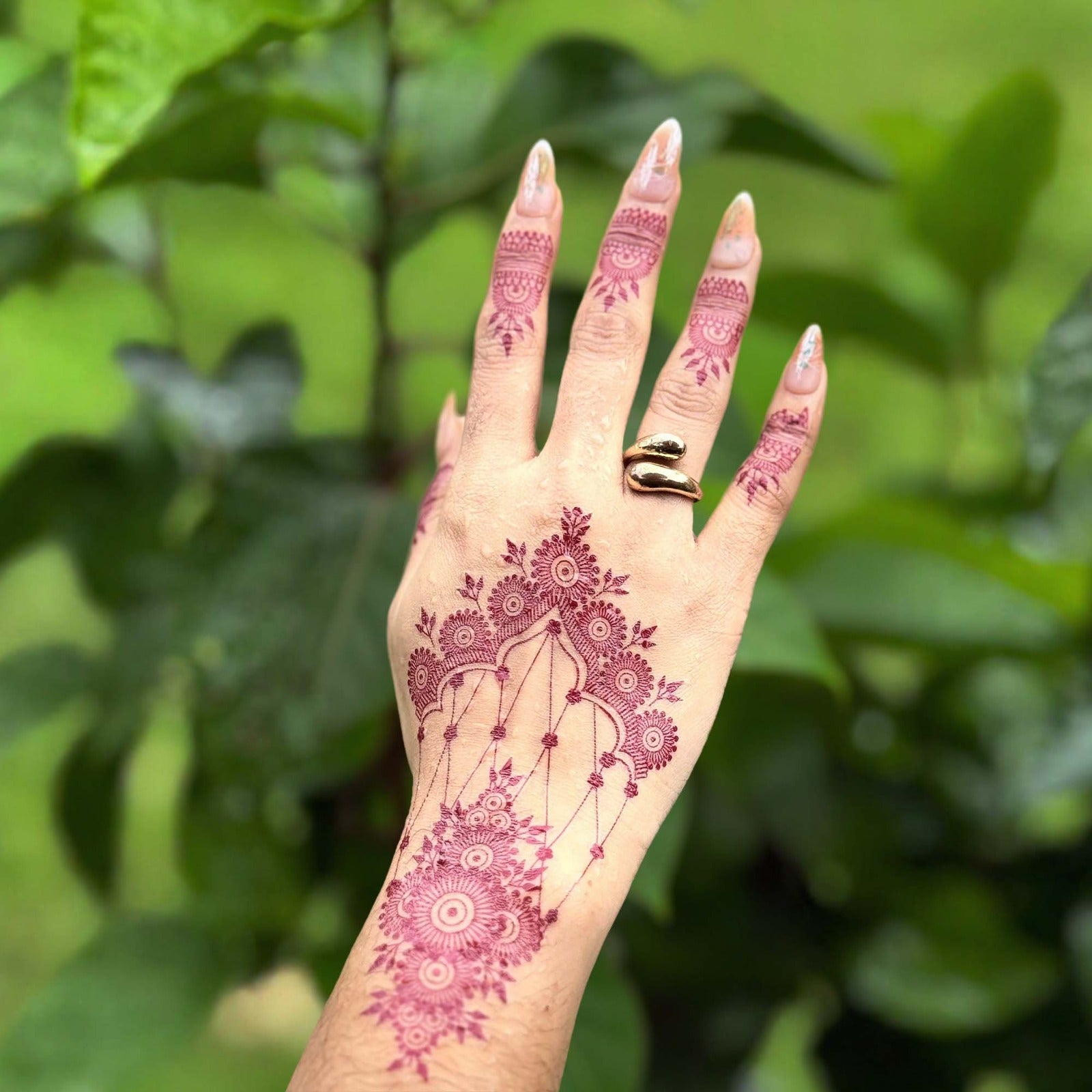 Hand with pink henna design against a green leafy background