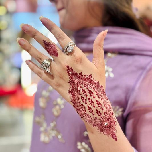 Hand with intricate red henna design, wearing multiple rings, against a blurred background.